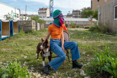 Fotoinszenierung: eine Person mit einer Mütze in Vogelform, welche den Kopf verdeckt, sitzt mit einem Hund auf einem Stuhl auf einer Stadtbrache