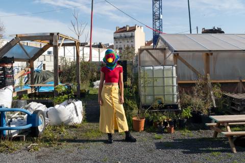Fotoinszenierung: eine Person mit einer Mütze in Vogelform, welche den Kopf verdeckt, steht zwischen Gewächshäusern und Gerätschaften eines urbanen Gartens