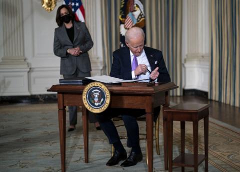 Joe Biden sitzt an einem Tisch im Weissen Haus, dahinter steht Kamala Harris
