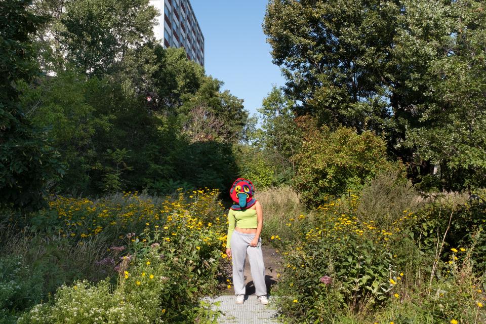 Fotoinszenierung: eine Person mit einer Mütze in Vogelform, welche den Kopf verdeckt, steht in einem urbanen Waldstück zwischen Sträuchern und Blumen
