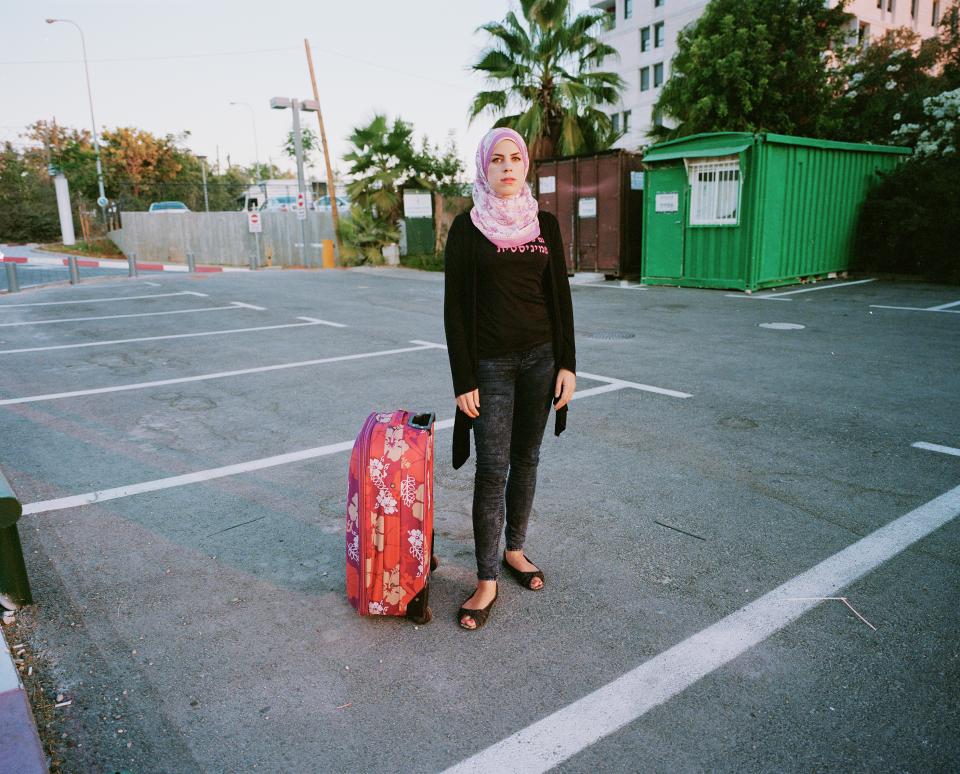 Aya steht mit ihrem Koffer auf einem Parkplatz